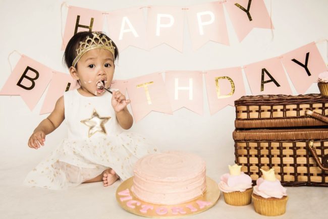 Baby Cake Smash Photography Singapore toddler young girl with a tiara eating cake with pin frosting with happy birthday banner behind her