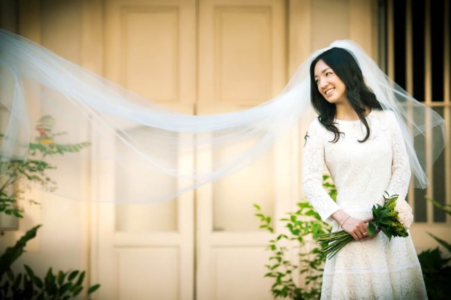 Bridal Photography Singapore bride standing outside shop house and holding flower bouquet with wedding veil blowing in the air