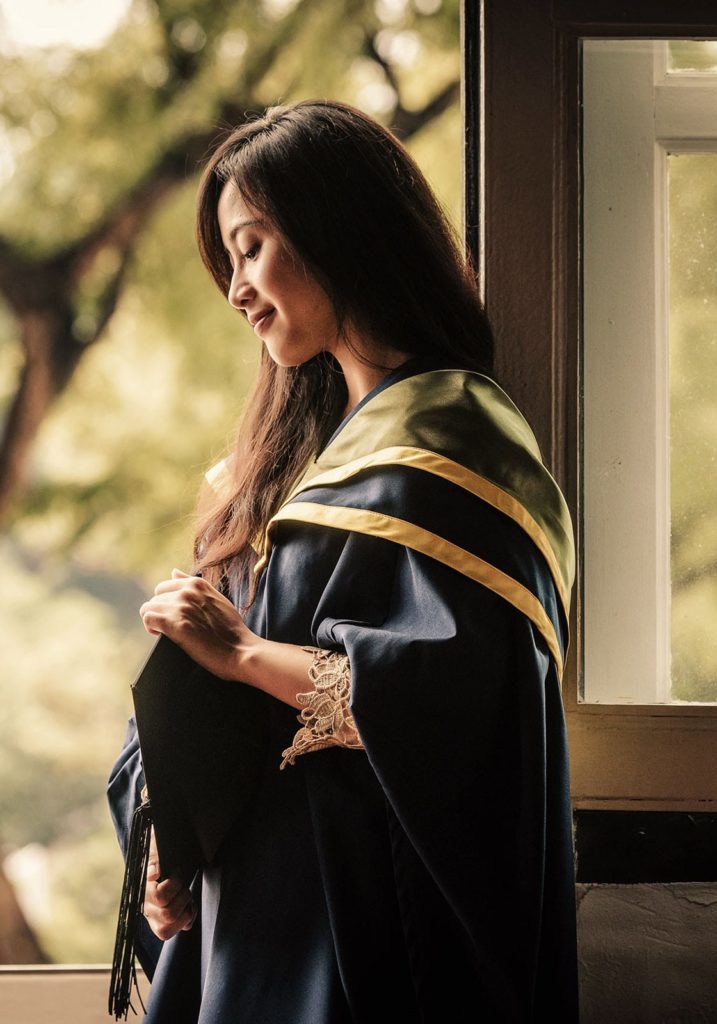 Graduation Pictures Singapore female graduate holding mortar board and leaning on door frame natural portrait, photographed at White Room Studio graduation photography session, 219 River Valley Road Singapore. Natural light family and graduation portrait 2026.