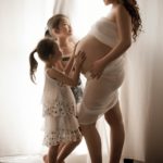 Maternity Photography Singapore 2 young girls touching pregnant mothers belly all standing next to white natural light curtain