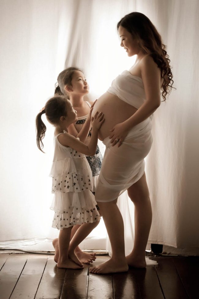 Maternity Photography Singapore 2 young girls touching pregnant mothers belly all standing next to white natural light curtain