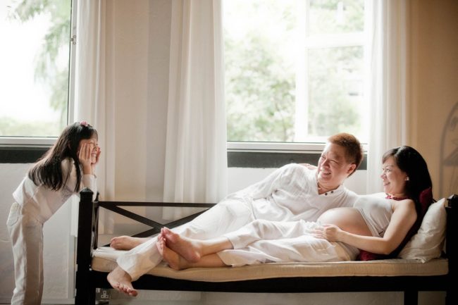 Maternity Photography Singapore family dressed in white clothes young girl child smiling at parents lying on sofa