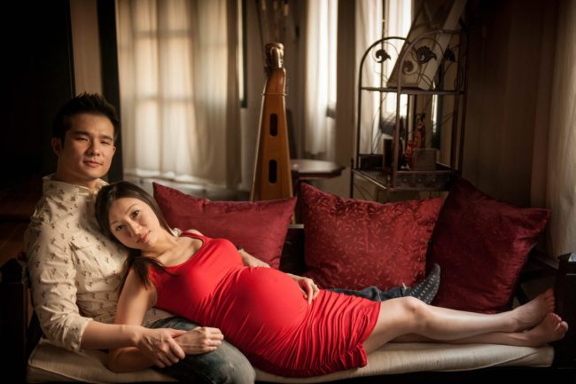Maternity Photoshoot Singapore pregnant wife in red dress leaning on husband on sofa with red cushions
