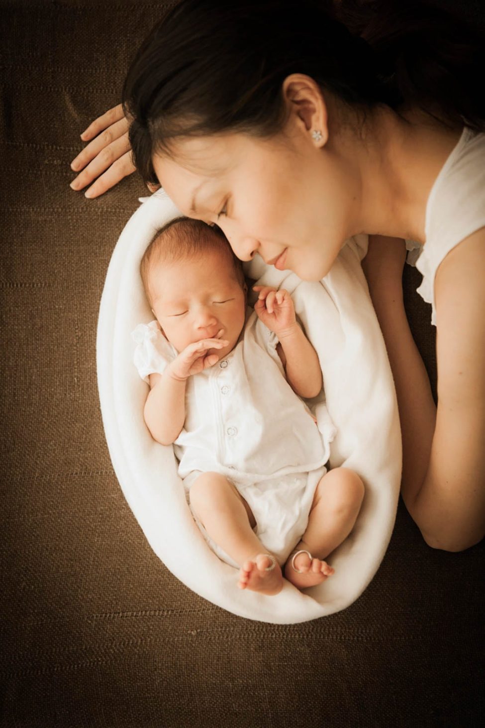 Newborn Photoshoot Singapore mother leaning near and looking at baby lying in white cushion, parents' wedding rings on baby's big toes