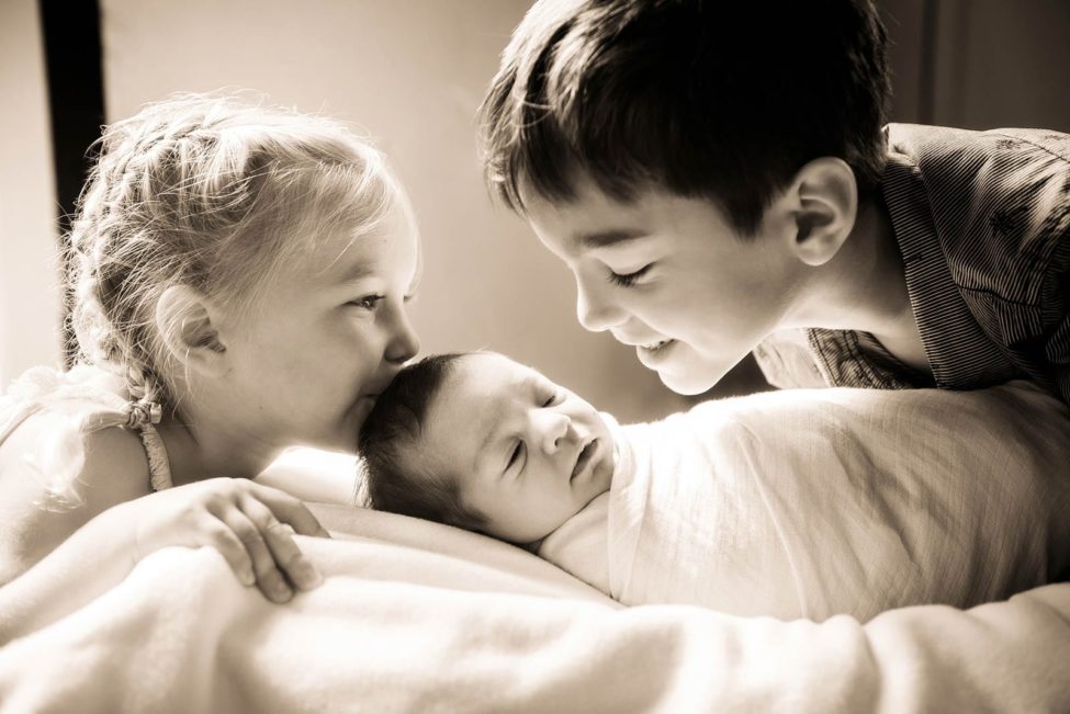Newborn Photoshoot Singapore 2 siblings close to and looking at baby sibling wrapped in white cloths for black and white portrait