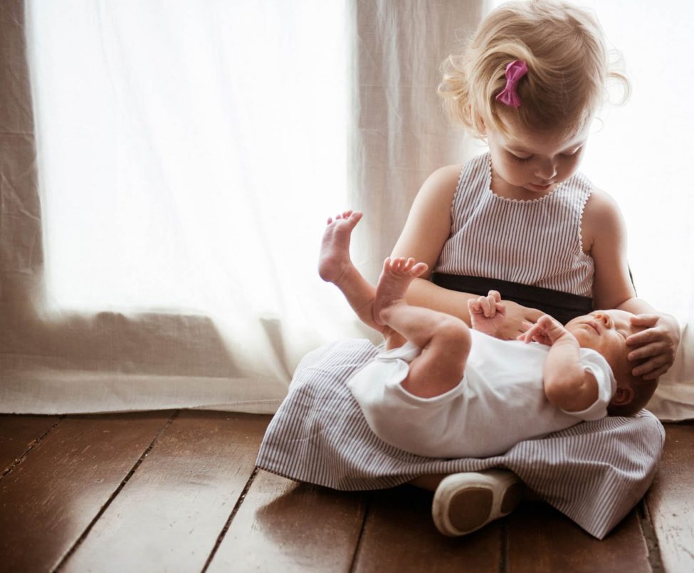 Newborn Photoshoot Singapore young girl sibling sitting on wooden floor and carrying baby sibling in lap natural light photography