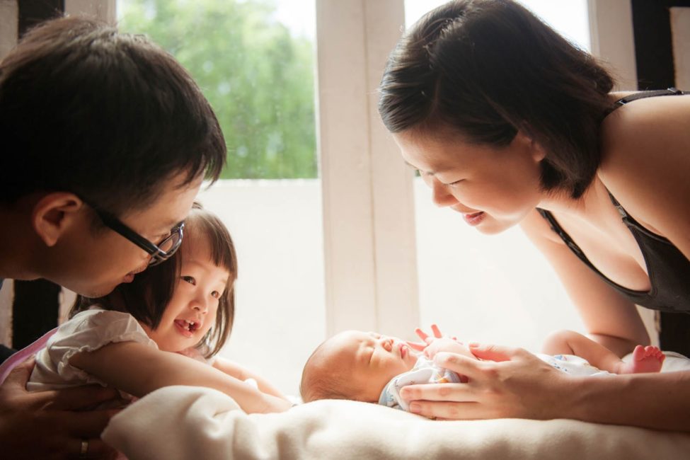 Newborn Shoot Singapore family members looking at baby sleeping parents and sibling smiling natural light photography