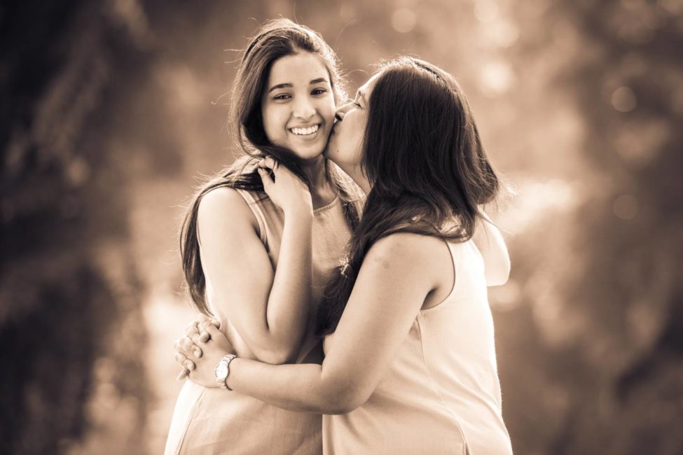 Generation photoshoot of mother hugging daughter's waist and kissing her cheek sepia tone portrait photography