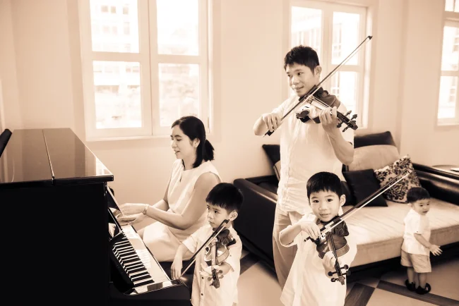 Family photoshoot package singapore of musically inclined family with parents and three young boys playing piano and violins, photographed by White Room Studio, 219 River Valley Road, Singapore. Natural light family portrait 2026