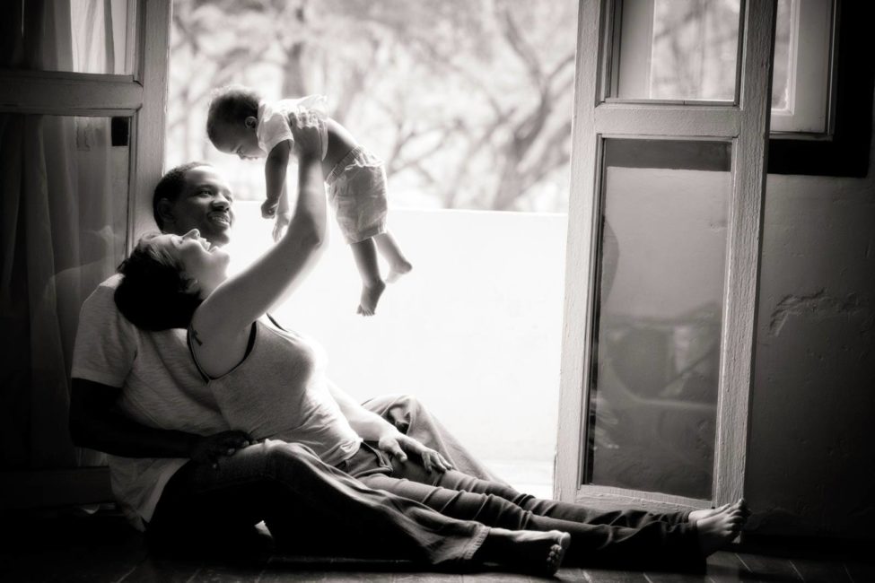 Professional family photoshoot with natural light wife lifting toddler above head and learning on husband against door frame black and white portrait photography