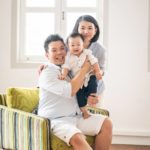 Professional family photoshoot Singapore father sitting on arm chair holding toddler child and mother standing posing for photos