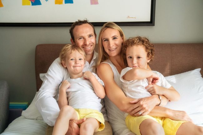 Home_photoshoot_Singapore_header Family photo of parents hugging their two young boys in white t-shirts, smiling at the camera and sitting on a bed