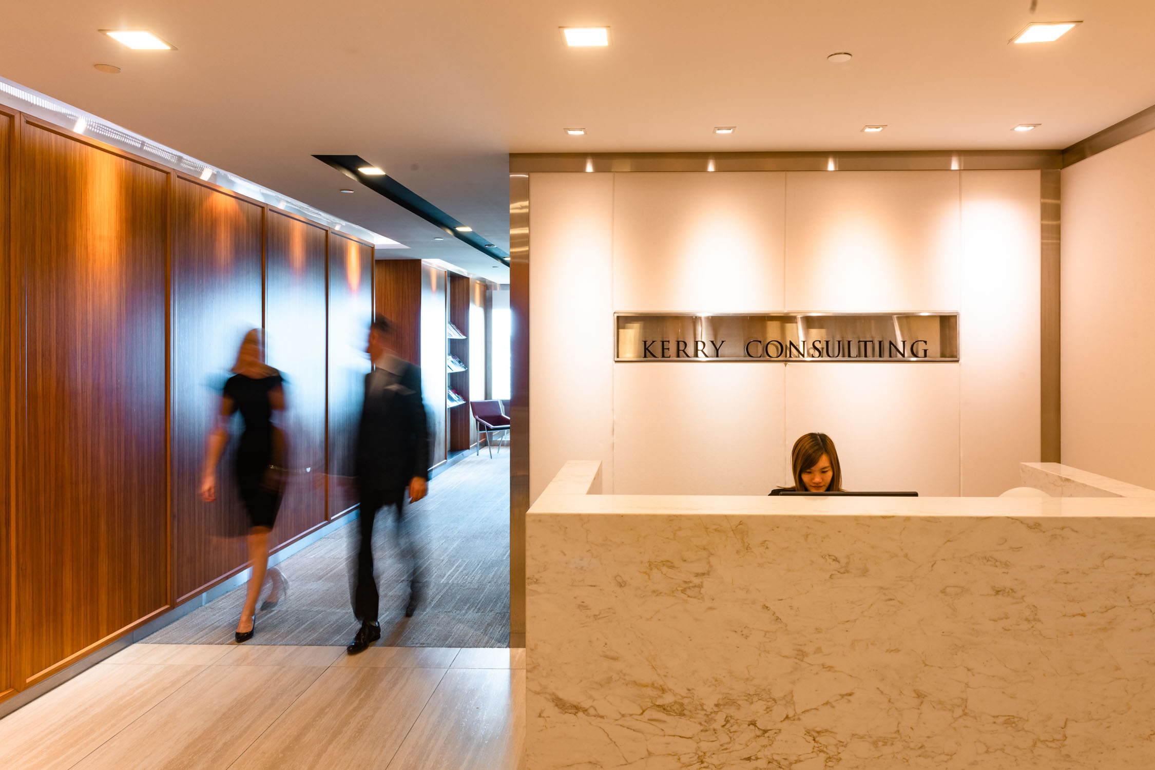 Long exposure shot of two people in business formal wear walking past company signage at reception area, a woman (receptionist) sitting down at the desk