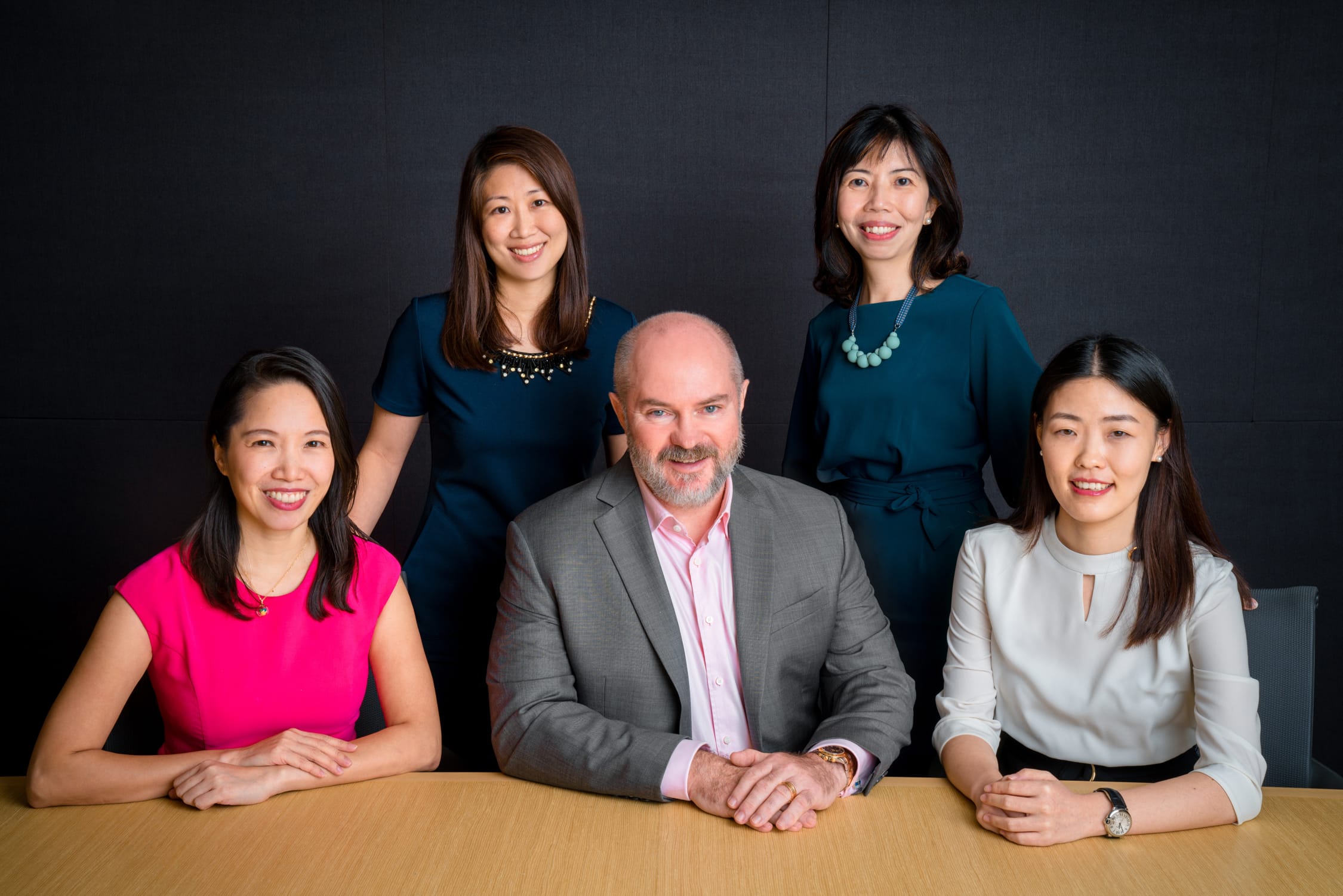 A group of male and female professionals smiling and sitting down at a desk posing for a corporate photoshoot in Singapore, White Room Studio
