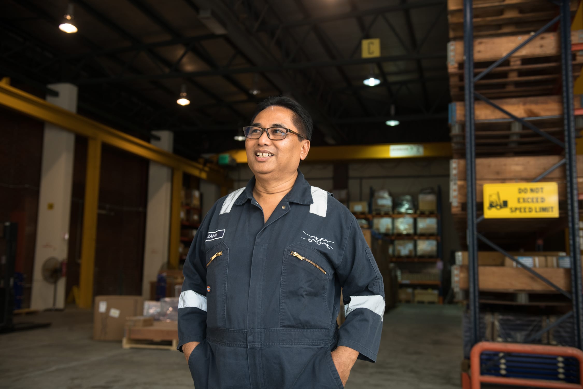 A man in a blue jumpsuit smiling for his headshot against a warehouse setting