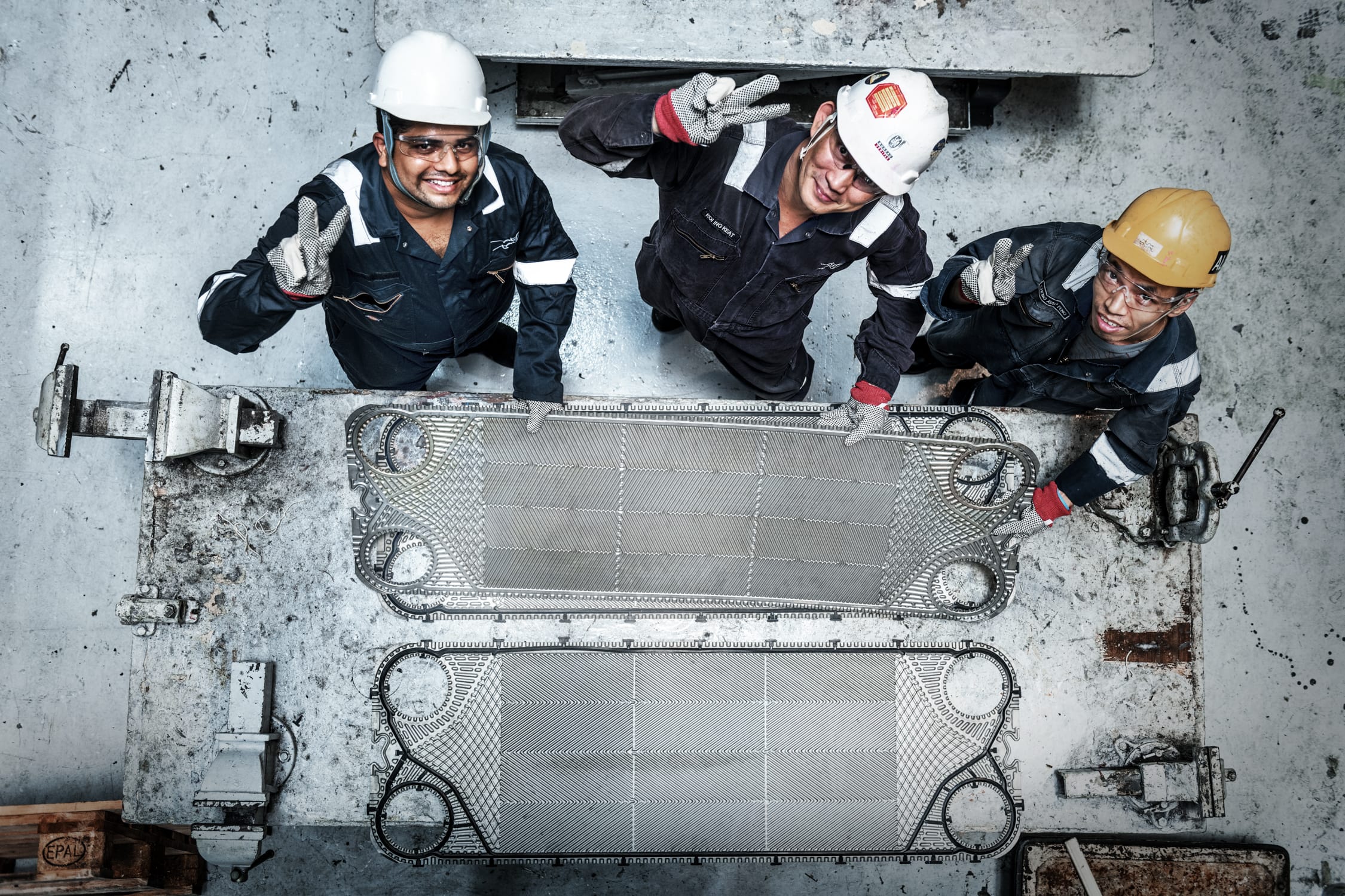 A top down photo of three workers wearing hard hats looking up and smiling with peace signs