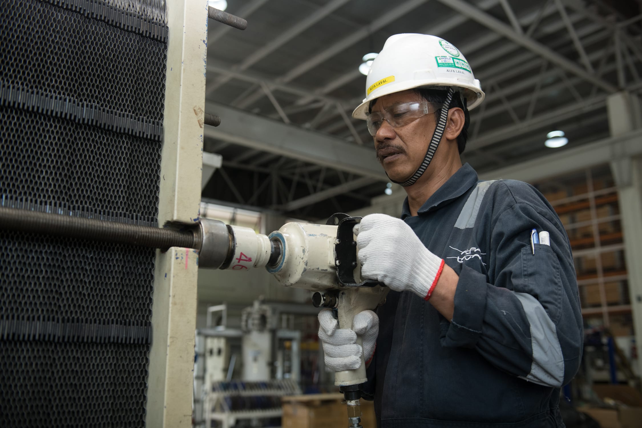 A worker wearing a white hard hat and blue jumpsuit and gloves uses a drilling machine