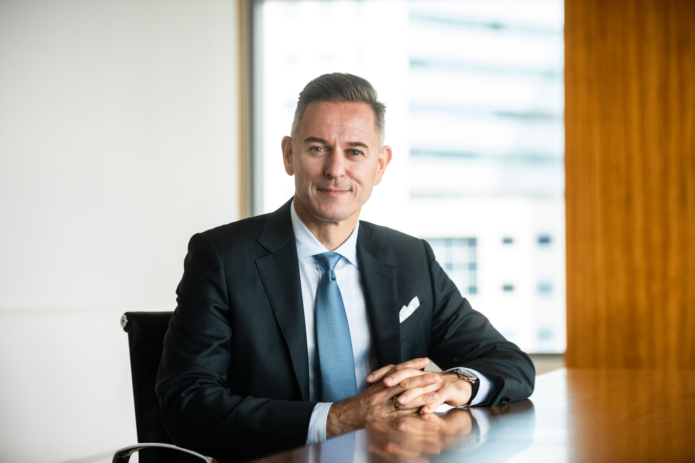 A man in a formal business suit and light blue tie smiles for his headshot in an office setting during a professional corporate photoshoot in Singapore