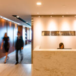 Long exposure shot of two people in business formal wear walking past company signage at reception area, a woman (receptionist) sitting down at the desk