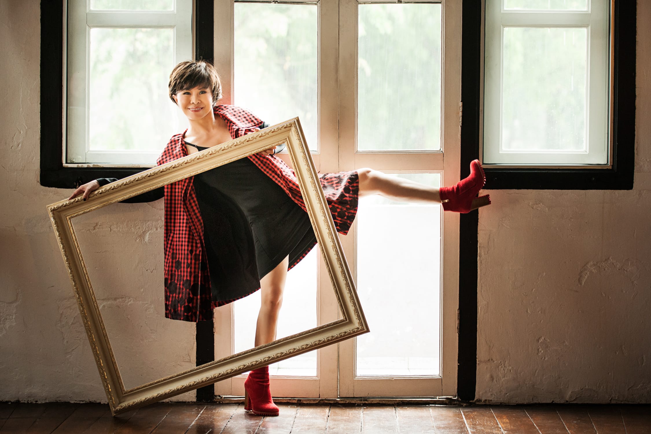 A woman in a black dress and pink coat poses for her headshot with one leg in the air while holding an empty picture frame during a professional indoor photo shoot in Singapore