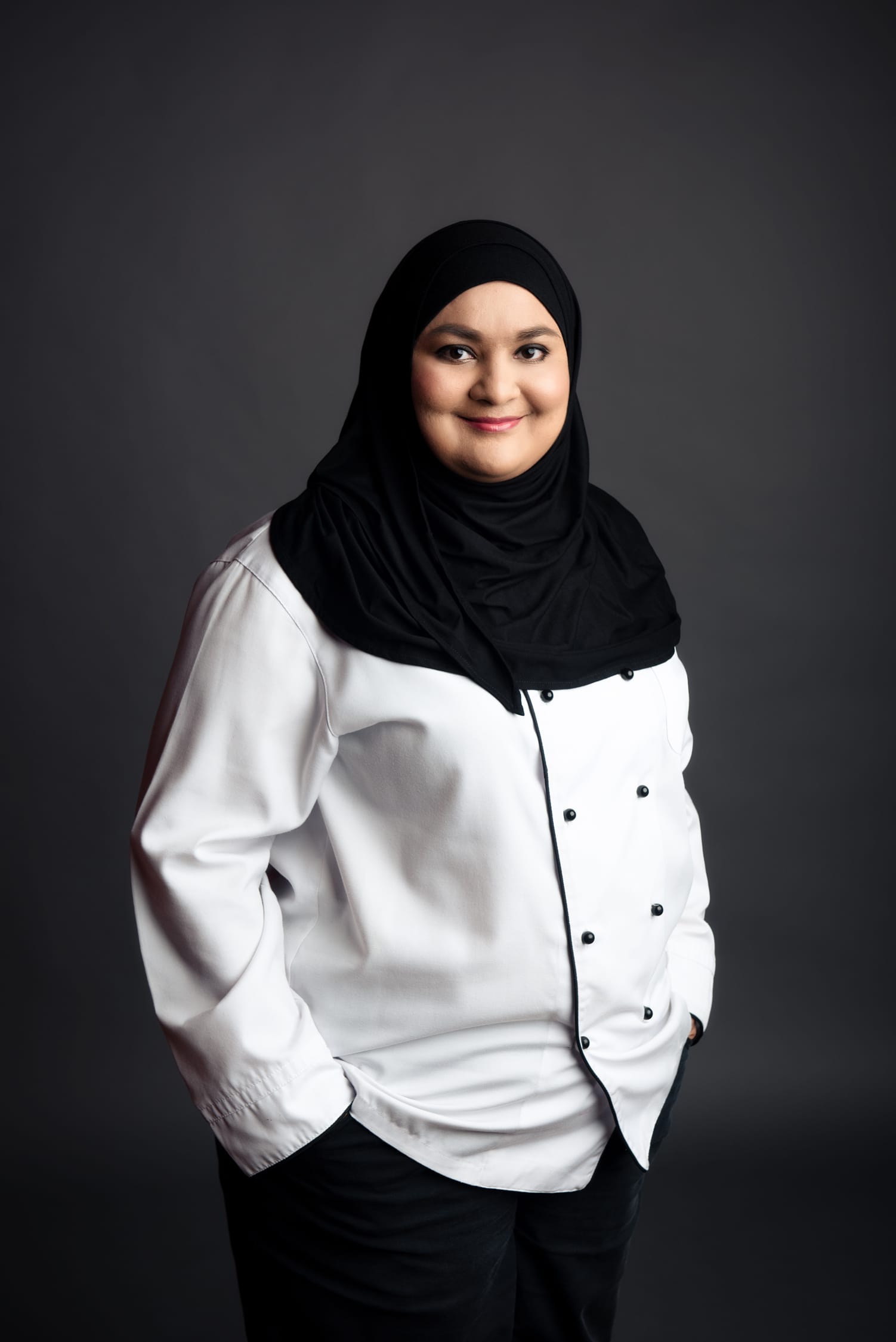 A woman wearing a hijab and chef's uniform poses for her headshot against a black studio background during a professional indoor photo shoot in Singapore