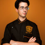 A man in a chef's uniform (TWG Tea) poses for his headshot with his arms folded across his chest, standing against an orange background during a professional indoor photo shoot in Singapore