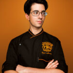 A man in a chef's uniform (TWG Tea) poses for his headshot with his arms folded across his chest, standing against an orange background during a professional branding photoshoot in Singapore Credit: White Room Studio