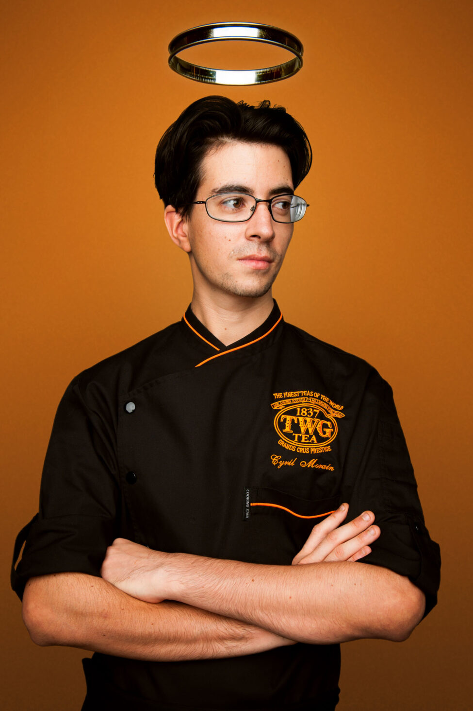 A man in a chef's uniform (TWG Tea) poses for his headshot with his arms folded across his chest, standing against an orange background during a professional branding photoshoot in Singapore Credit: White Room Studio