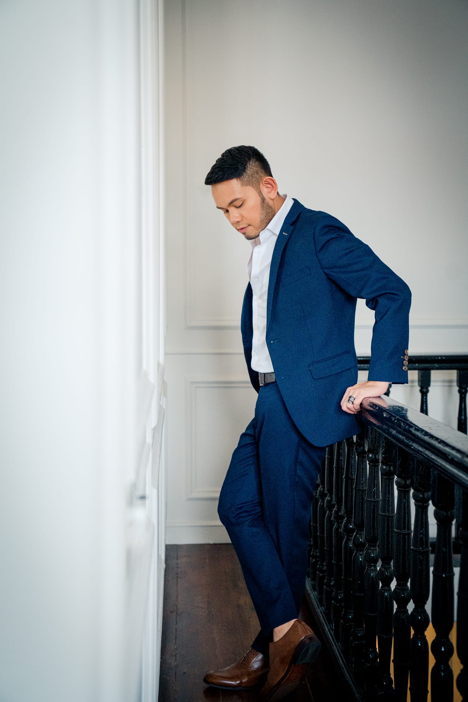 A man in a blue blazer and suit and brown leather shoes poses against a staircase railing during a professional indoor photo shoot in Singapore