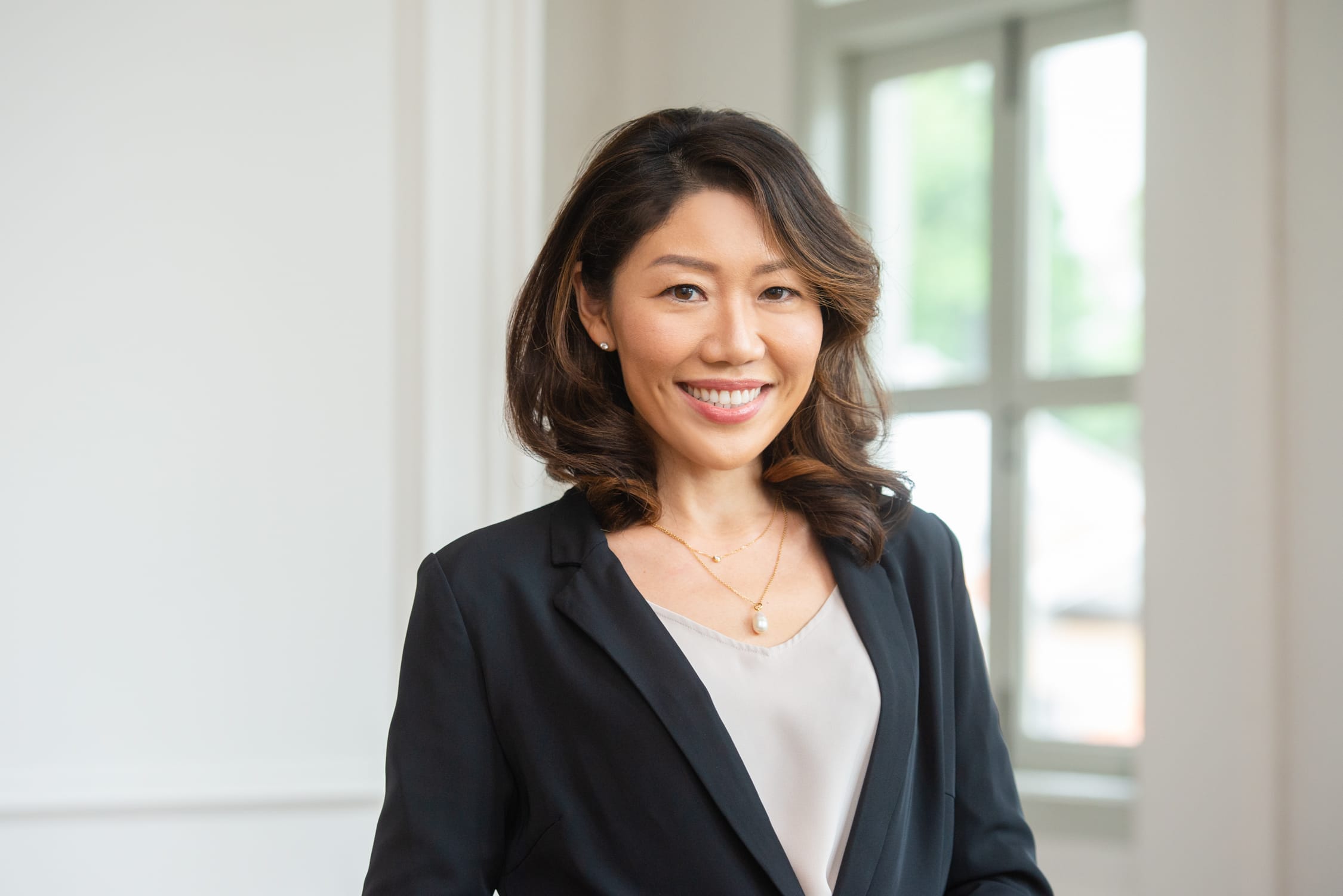 A woman in a black blazer and light grey blouse poses for her headshot with tall and large windows in the background