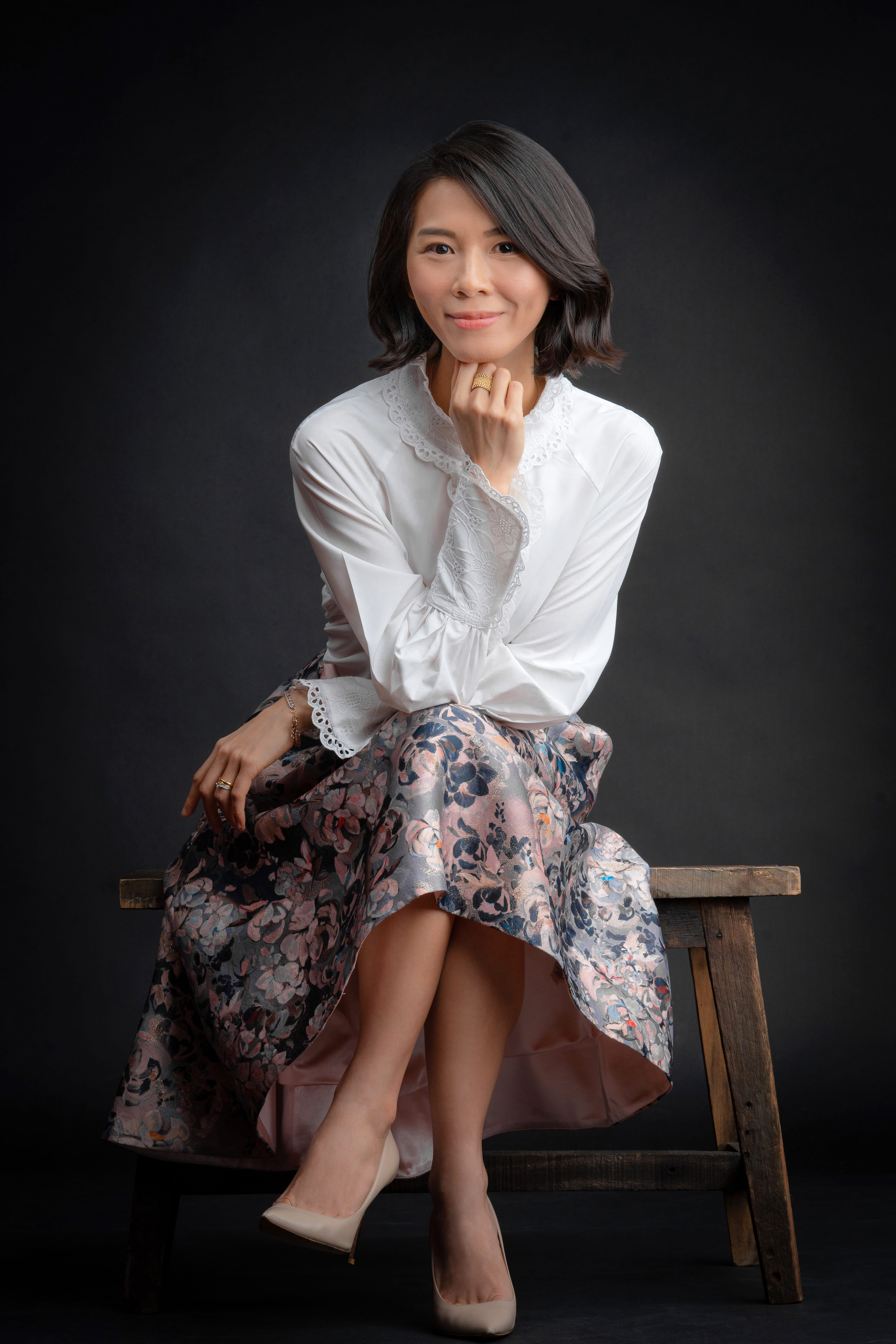 A woman in a white blouse and patterned skirt sitting on a wooden chair posing for a headshot against a black studio background during a corporate photoshoot in Singapore Credit: White Room Studio
