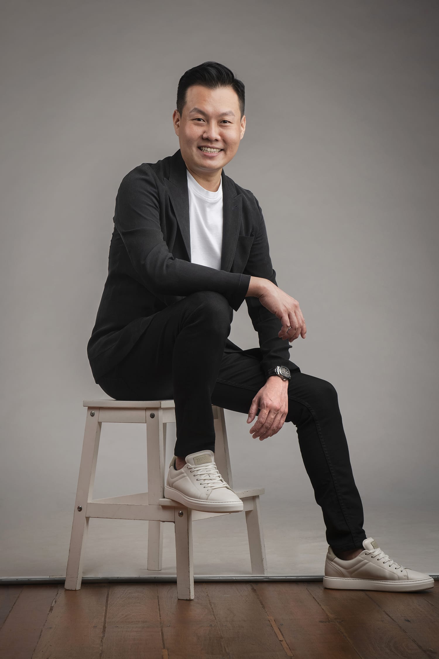 A man wearing black blazer and pants poses for his headshot while sitting on a stool against a grey studio background during a corporate photoshoot in Singapore Credit: White Room Studio