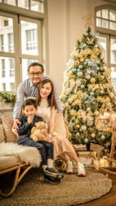 Mother and father with son hugging teddy bear with Christmas tree in the background during a Christmas family photoshoot in Singapore Credit: White Room Studio