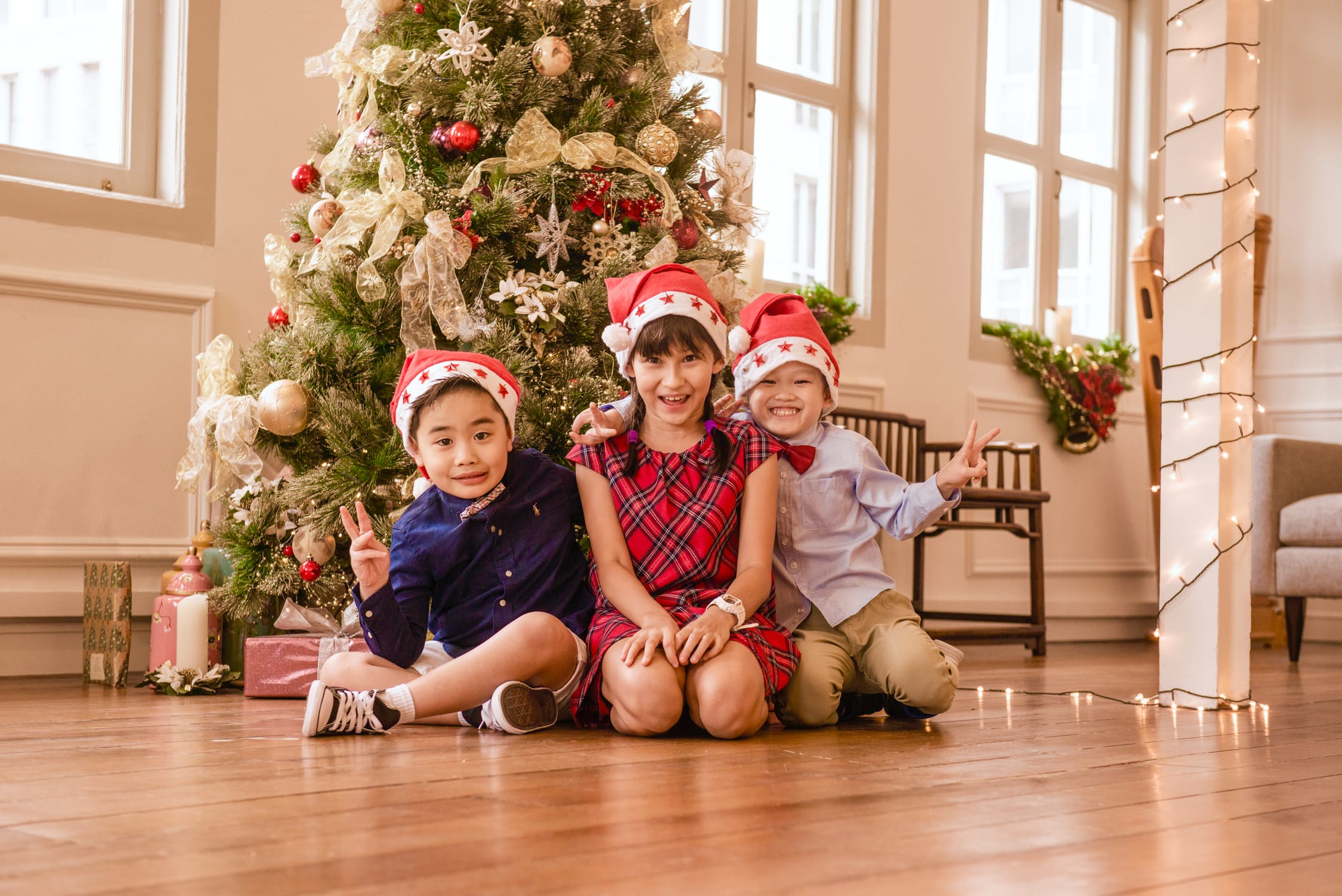 Three young children smiling with Christmas tree and fairy lights during family Christmas photoshoot in Singapore Credit: White Room Studio