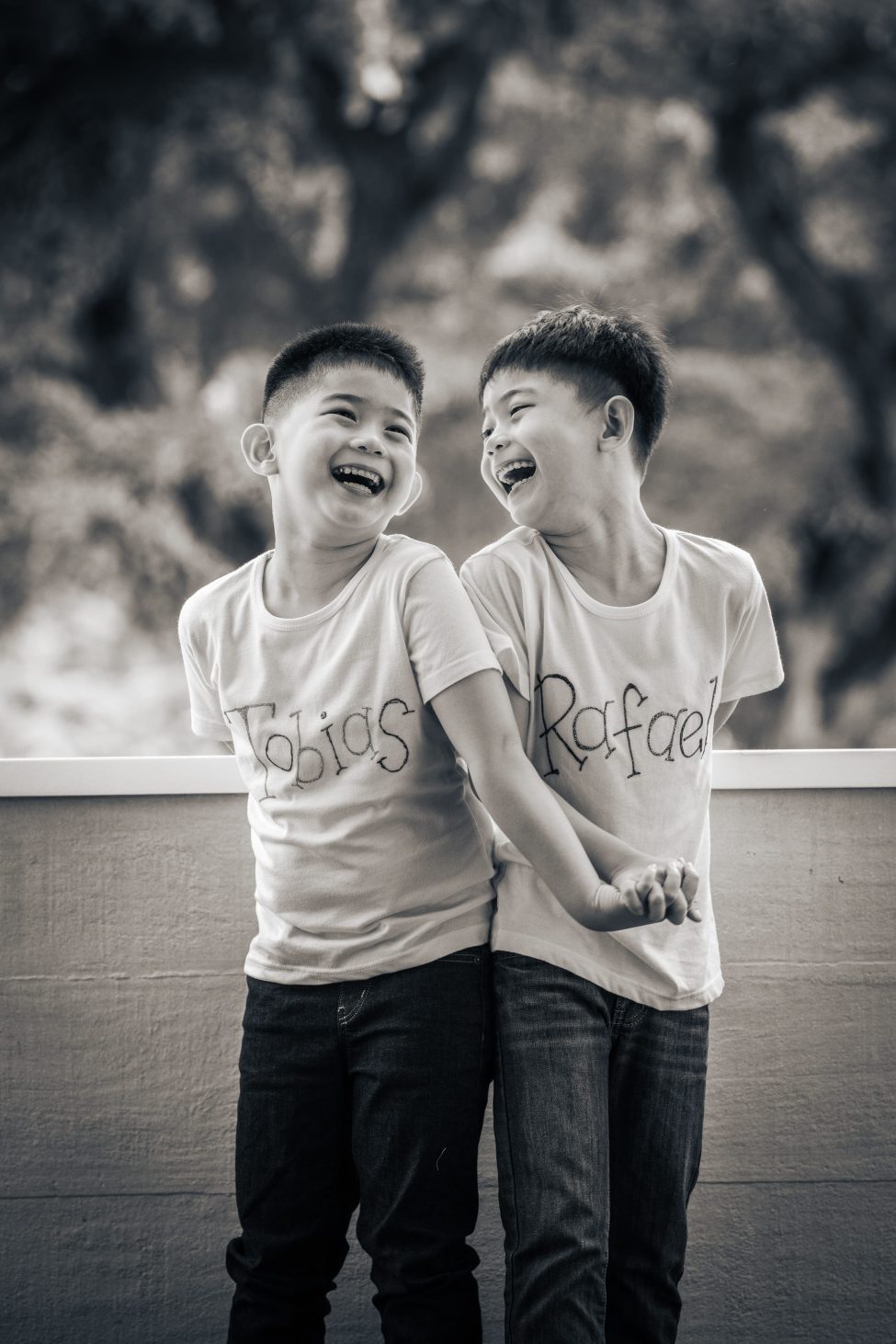 Black and white photo of two boys brothers in white t-shirt and jeans holding hands smiling at each other during a K2 kindergarten graduation photoshoot in Singapore Credit: White Room Studio