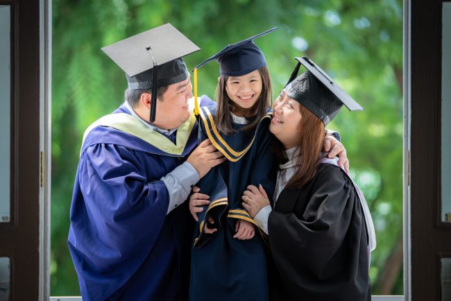 Smiling Father and Mother in university graduation gown carrying daughter in K2 kindergarten graduation gown and cap with nature scenery background during a k2 kindergarten graduation photoshoot in Singapore Credit: White Room Credit Studio