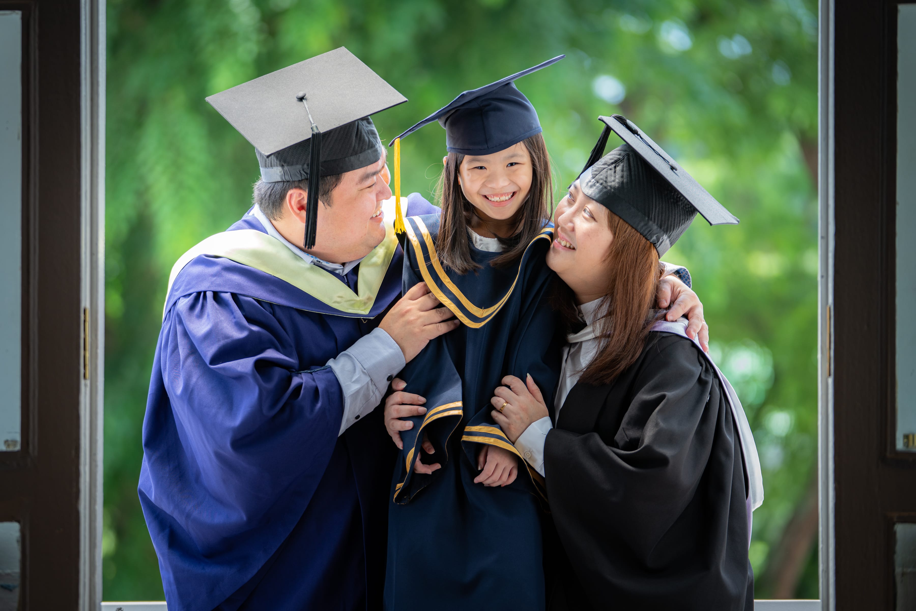 Smiling Father and Mother in university graduation gown carrying daughter in K2 kindergarten graduation gown and cap with nature scenery background during a k2 kindergarten graduation photoshoot in Singapore Credit: White Room Credit Studio