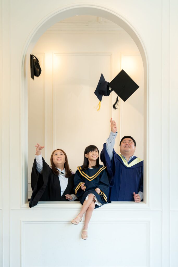 Smiling Father and Mother in university graduation gown carrying daughter in K2 kindergarten graduation gown and cap during a k2 graduation photoshoot in Singapore Credit: White Room Credit Studio