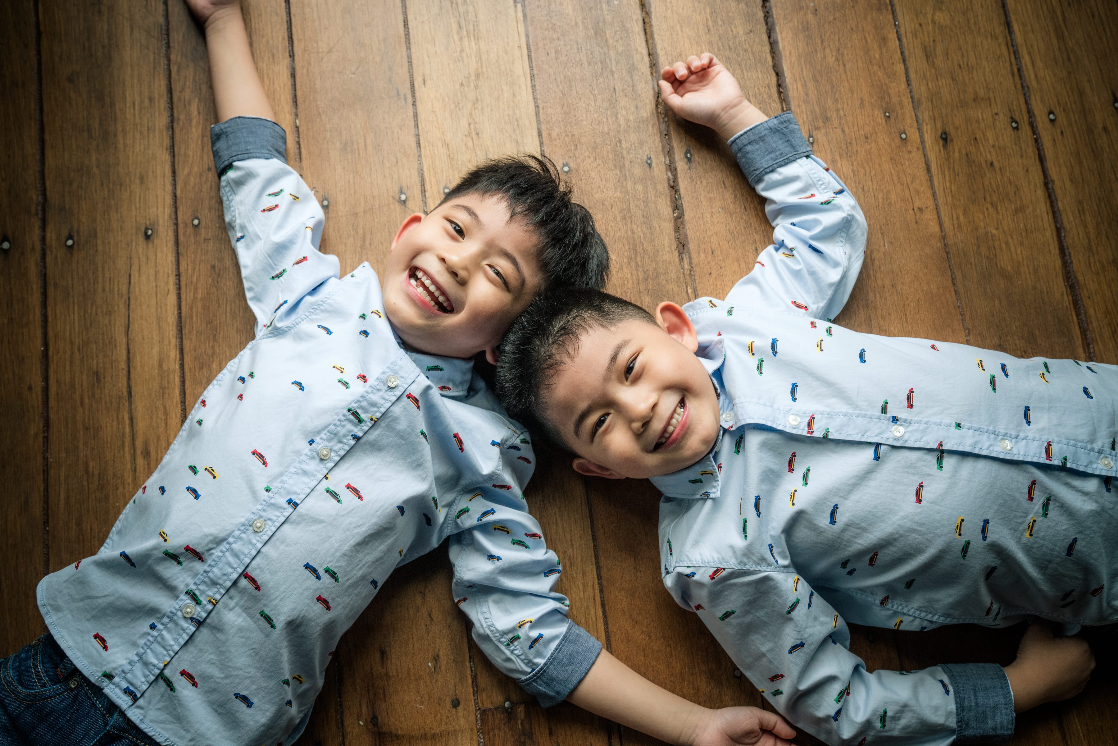 Two boys brothers in matching shirts lying on hardwood floors smiling at the camera during k2 kindergarten graduation shoot in Singapore Credit: White Room Studio