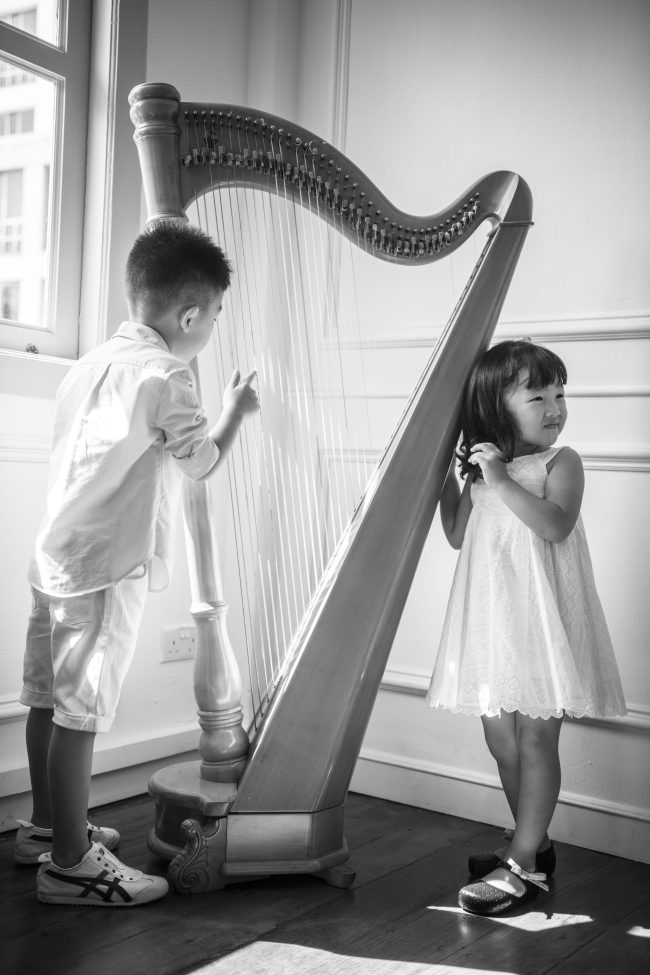Black and white photo of two young kids playing with harp near windows with natural light during a family photoshoot in Singapore Credit: White Room Studio