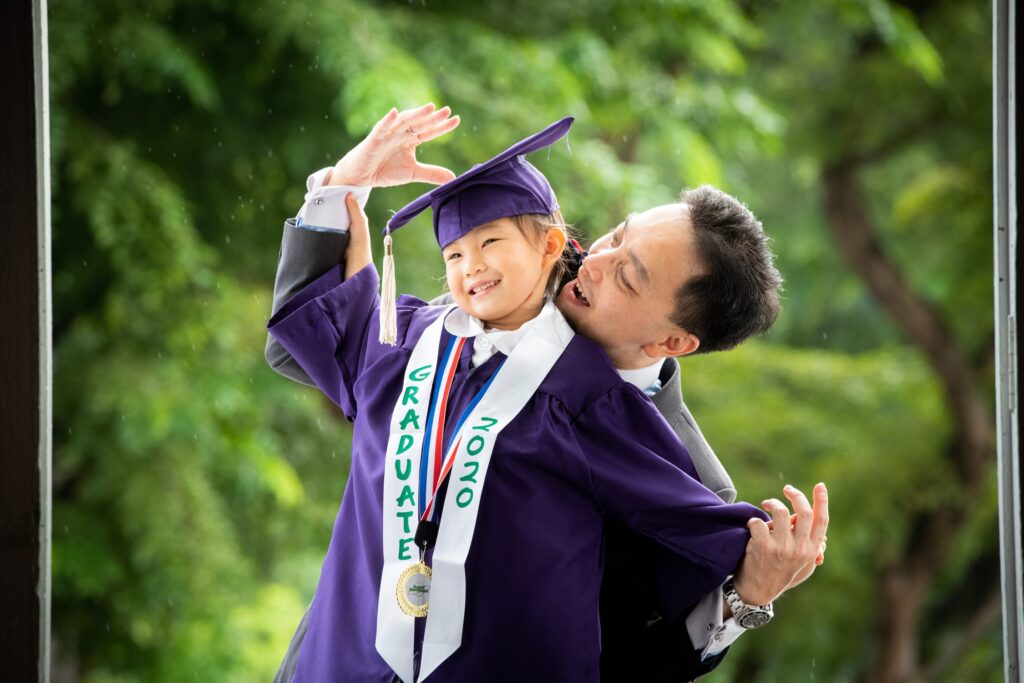 kindergarten-graduation-k2-photoshoot-singapore-white-room-studio Smiling Father and daughter in K2 kindergarten graduation gown and cap with nature scenery background during a kindergarten graduation k2 photoshoot in Singapore Credit: White Room Studio