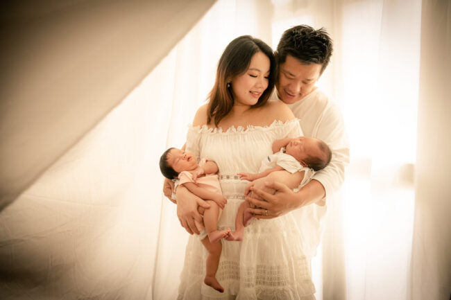 Professional Family & Newborn Photoshoot in Photography Studio, Singapore Mother and father carrying their newborn baby twins in a photography studio setting during a natural light newborn photoshoot in Singapore, White Room Studio. Credit: White Room Studio Pte Ltd