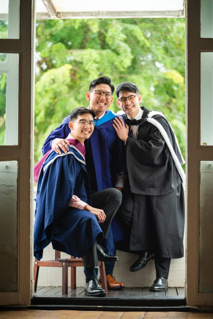 Three siblings (brothers) in graduation gowns smiling in a photography studio setting during a graduation photoshoot in Singapore, White Room Studio. Credit: White Room Studio Pte Ltd