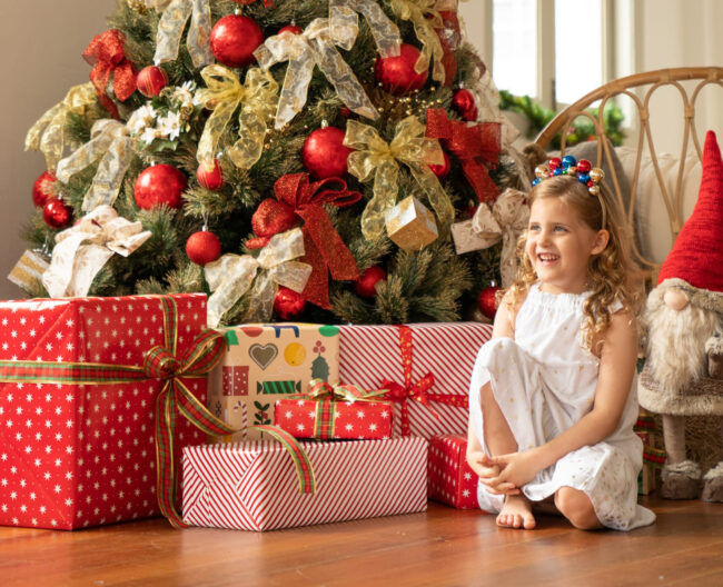 Smiling girl with Christmas tree and Christmas presents in a photography studio setting during a Christmas family photoshoot in Singapore, White Room Studio. Credit: White Room Studio Pte Ltd