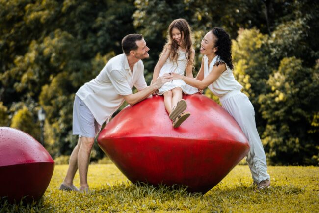 Mother and father and daughter smiling at each other during an outdoor family photoshoot in Singapore, Botanic Gardens. Credit: White Room Studio Pte Ltd