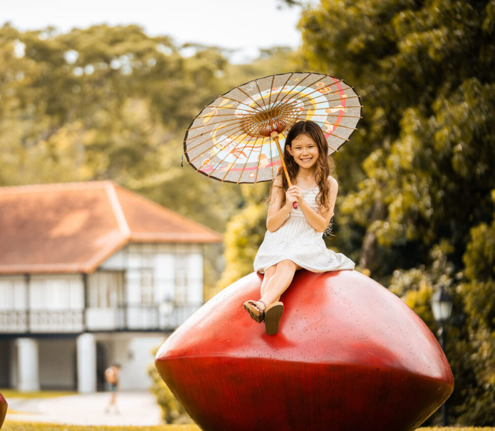 Girl carrying umbrella sitting on saga seed during an outdoor family photoshoot in Singapore, Botanic Gardens. Credit: White Room Studio Pte Ltd