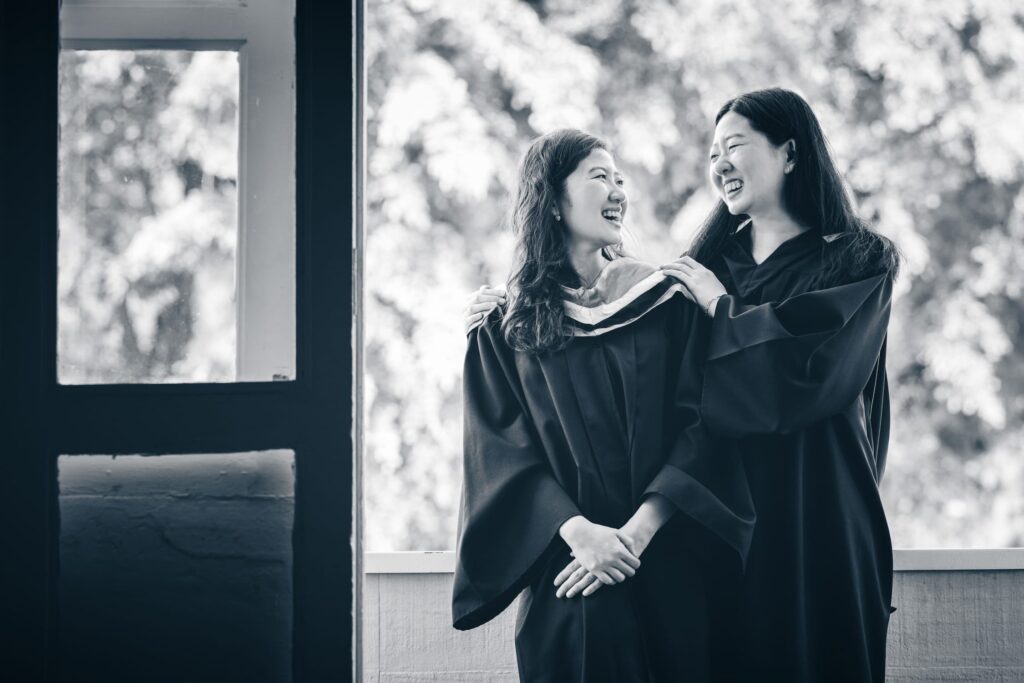 Professional Family and Graduation Photoshoot in Photography Studio, Singapore Two sisters in graduation gown smiling at each other in a photography studio setting during a graduation photoshoot in Singapore, White Room Studio. Credit: White Room Studio Pte Ltd