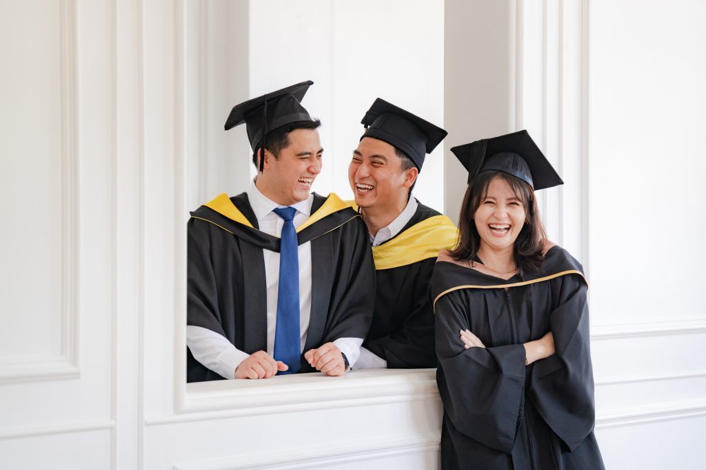 Three graduates in caps and gowns smile and laugh together near a white wall indoors, capturing the joy of achievement—perfect for a Professional Family Photoshoot in a Photography Studio in Singapore.