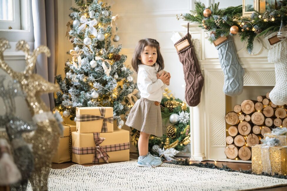 Young girl stands by a Christmas tree and fireplace with stockings, surrounded by presents and festive decor during a Professional Family Photoshoot in Photography Studio, White Room Studio Singapore.